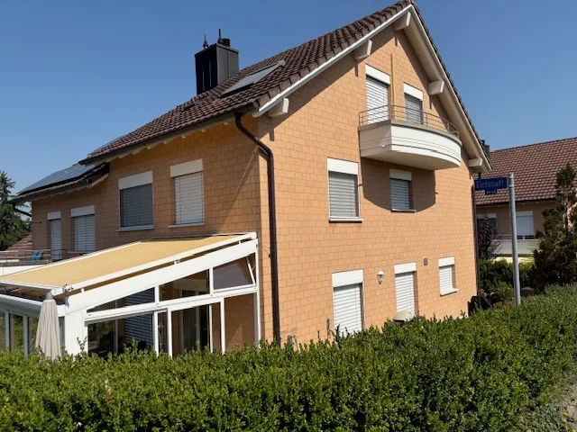A warm orange multi-story residential house with solar panels, balcony, and lush green hedges under blue sky.