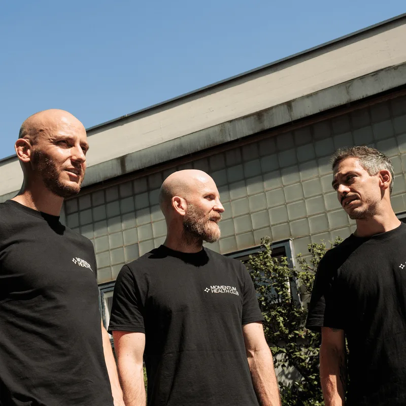 Three founders of MOMENTUM Health Club stand together outdoors wearing matching black branded shirts.