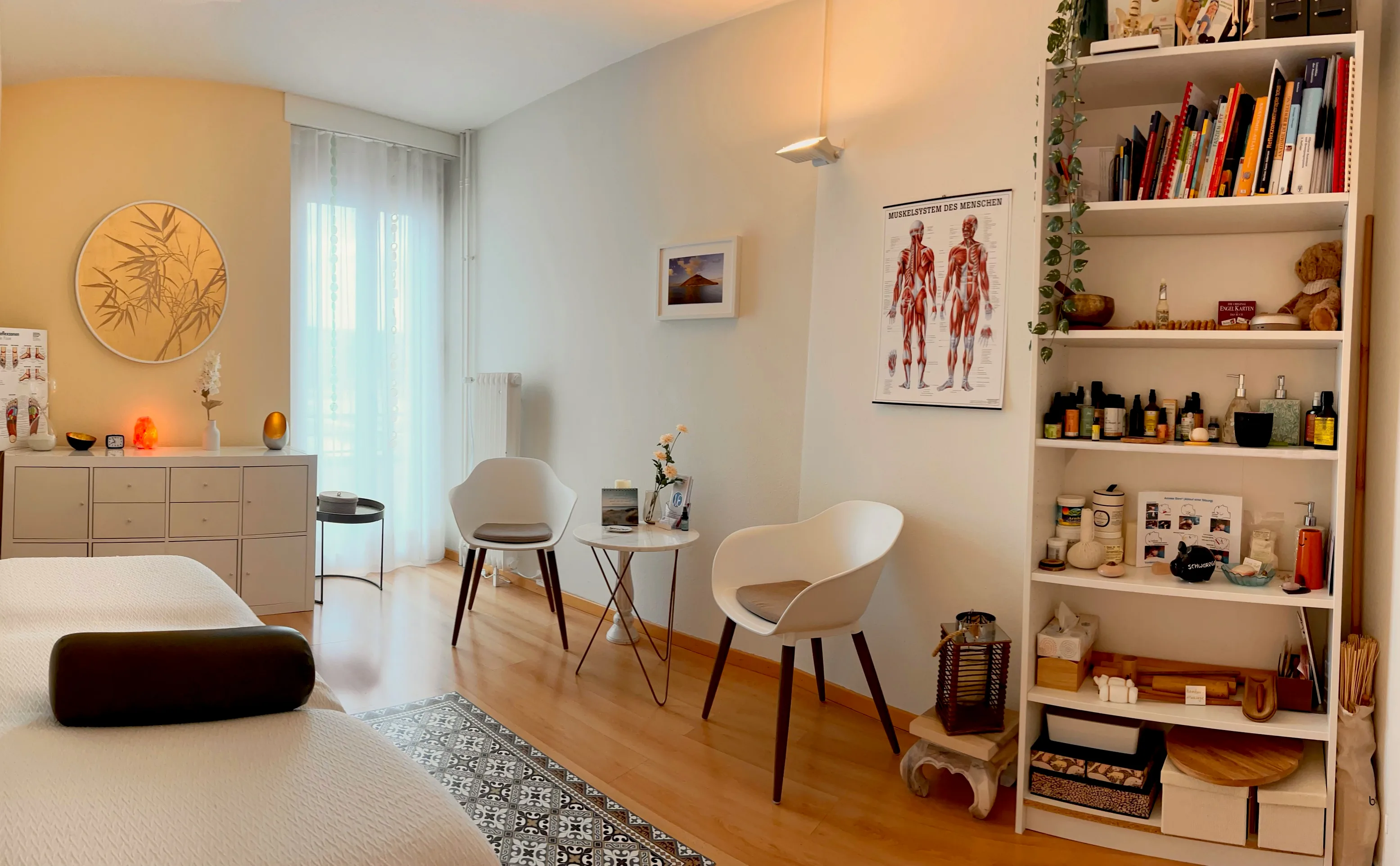 Bright welcoming massage and therapy practice room with treatment table, shelves of oils, and anatomical posters.