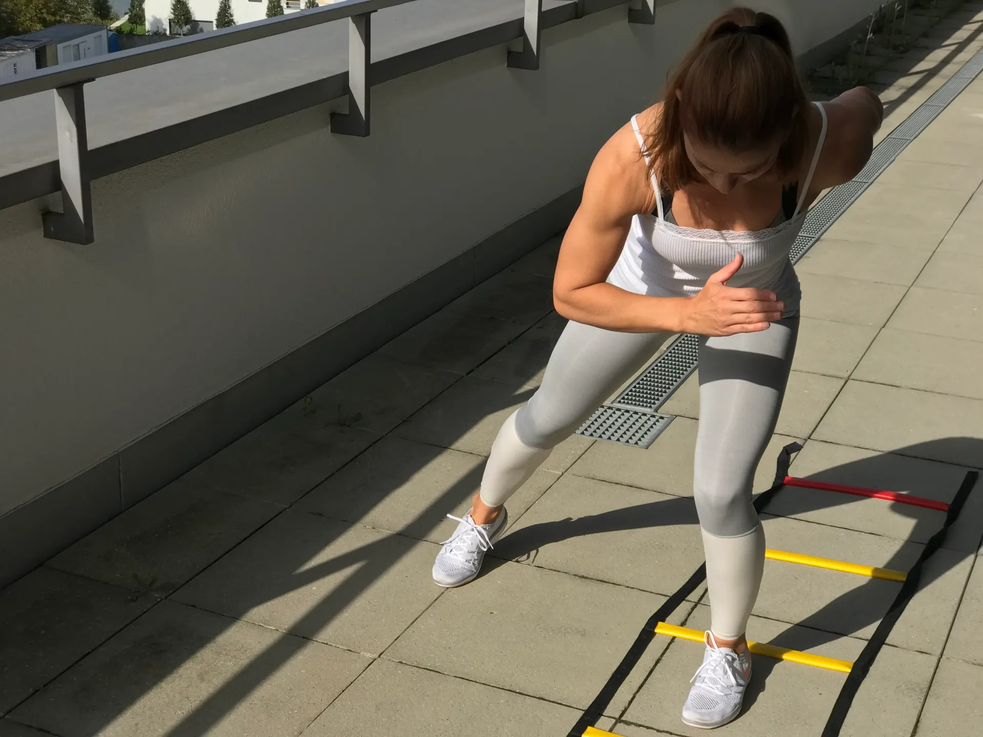 A woman performs a lateral agility ladder drill on a sunny rooftop terrace.
