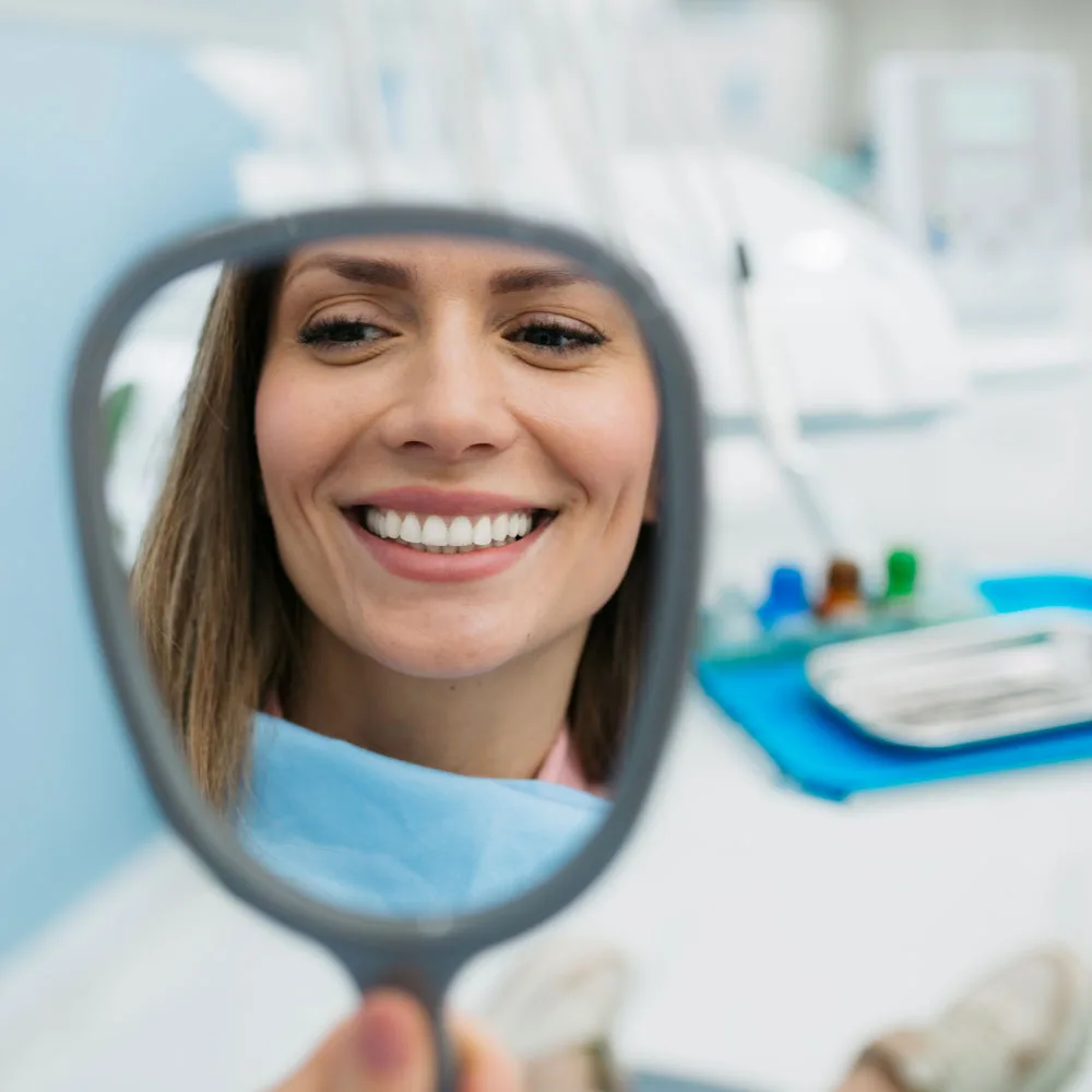 Young woman admiring her smile in a dental mirror, illustrating the emotional moment of regained confidence after prosthetic treatment.