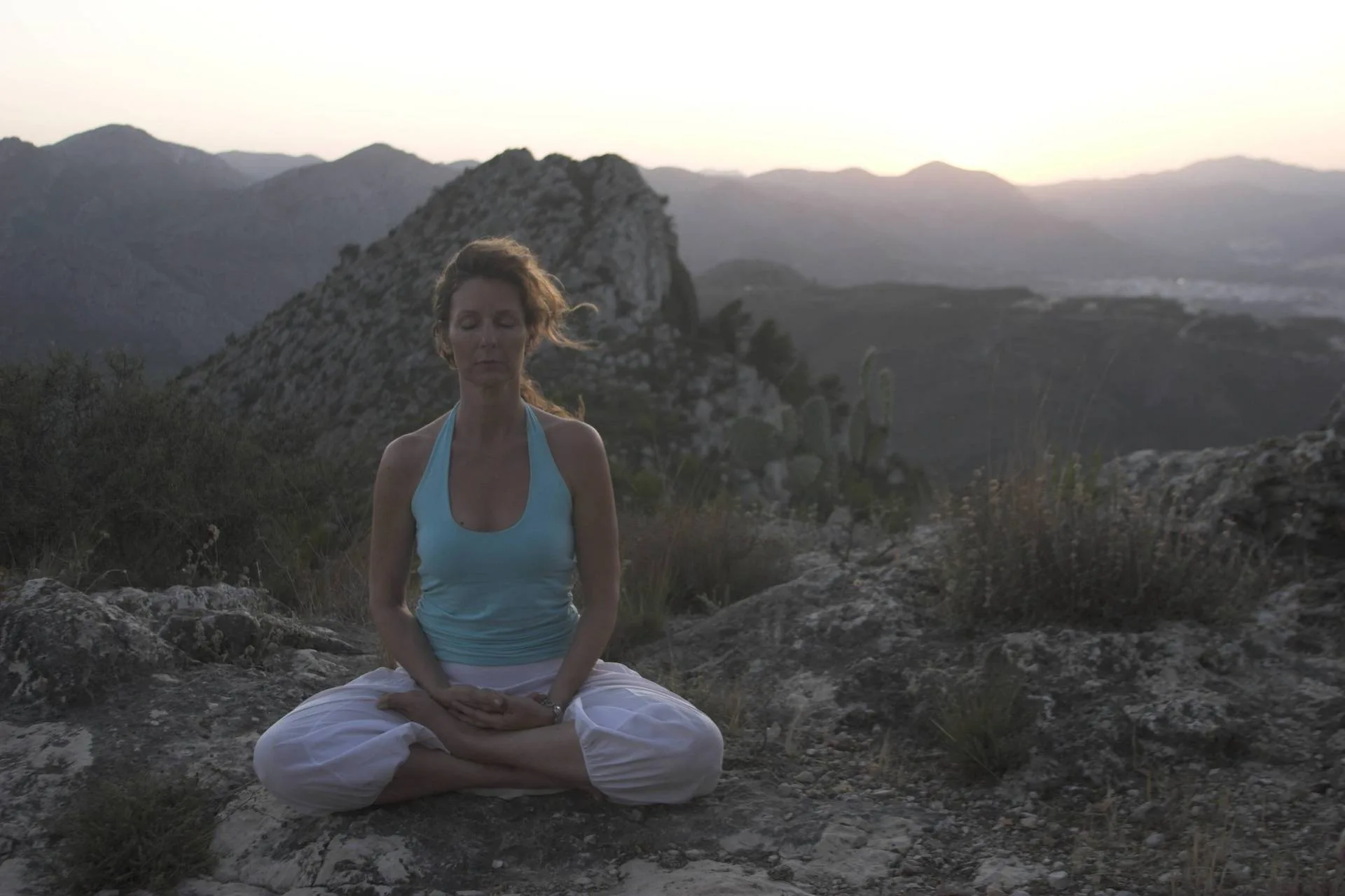 A woman meditates serenely in lotus pose on a rocky mountain summit at dusk.