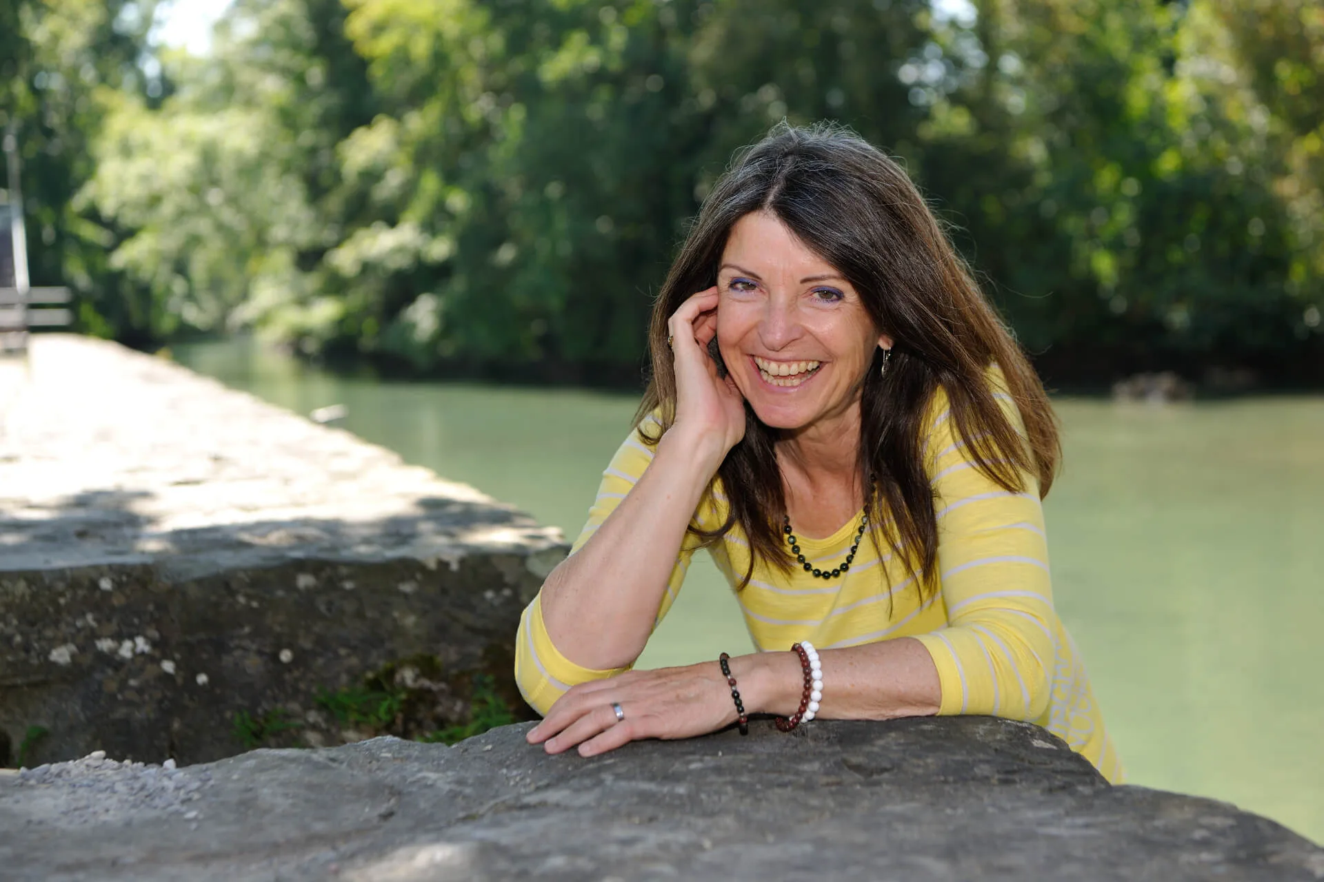 Ursula Rothenfluh smiling warmly by a riverside, conveying trust, approachability, and the authentic human connection at the heart of her coaching practice.