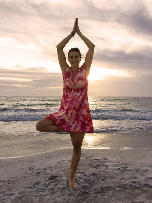 Woman in colorful dress performing tree yoga pose on a beach at sunset.