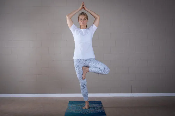 A woman balances gracefully in tree pose on a yoga mat in a studio.