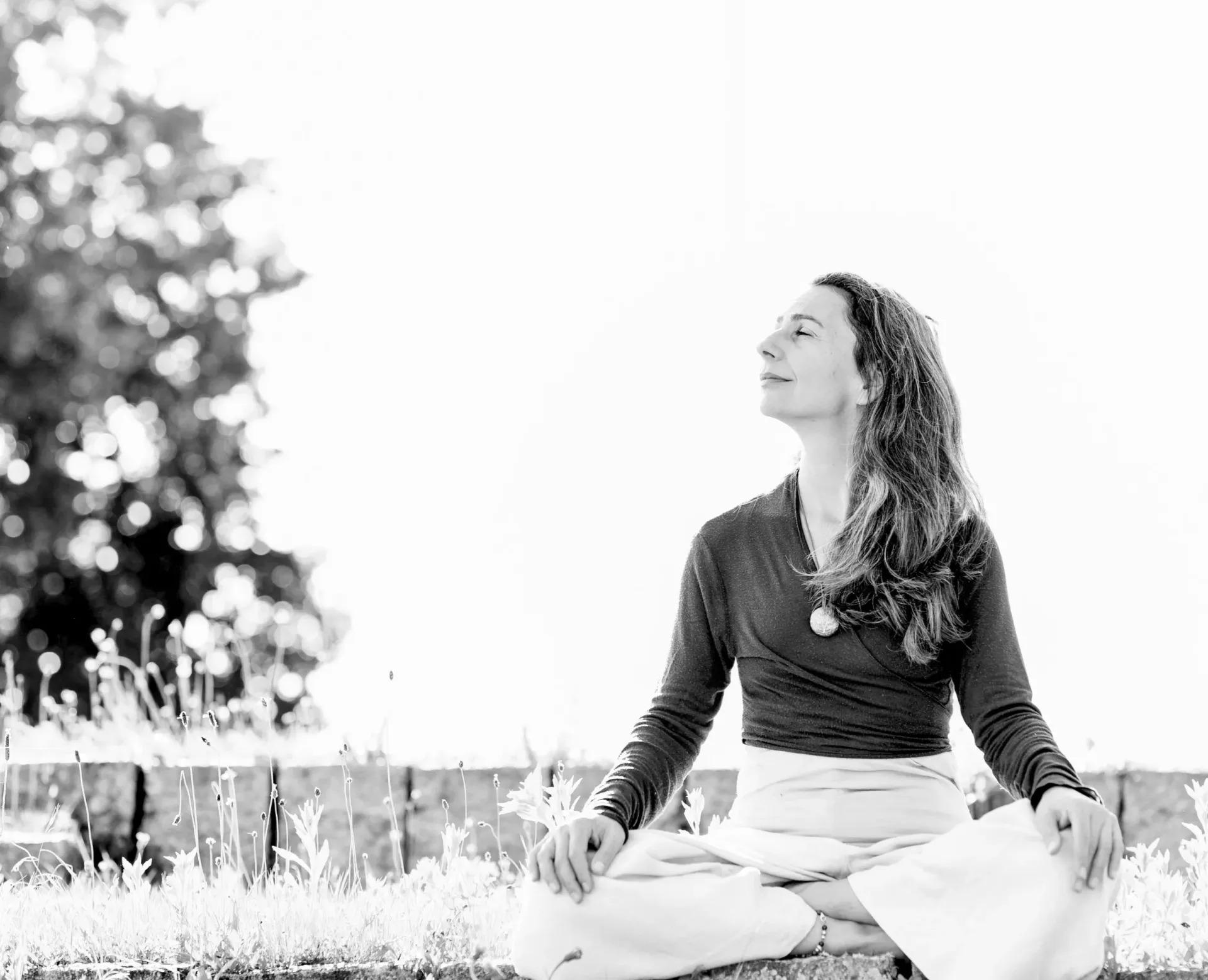 Romina Salerno sits peacefully in sunlit meadow grass, eyes closed — a serene portrait perfectly embodying body-mind presence and inner stillness.