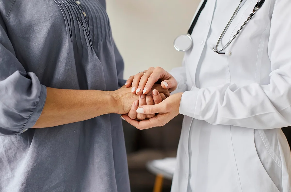 A doctor in a white coat gently holds a patient's hands in a compassionate gesture.