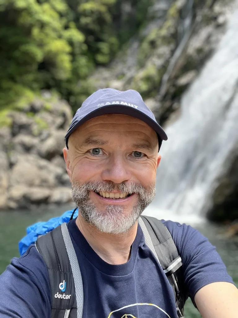 Male runner with hiking backpack smiling by waterfall, embodying the mountain running passion and outdoor adventure philosophy mentioned in article.