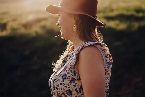 A woman in a floral dress and wide-brim hat stands serenely in golden sunlight outdoors.