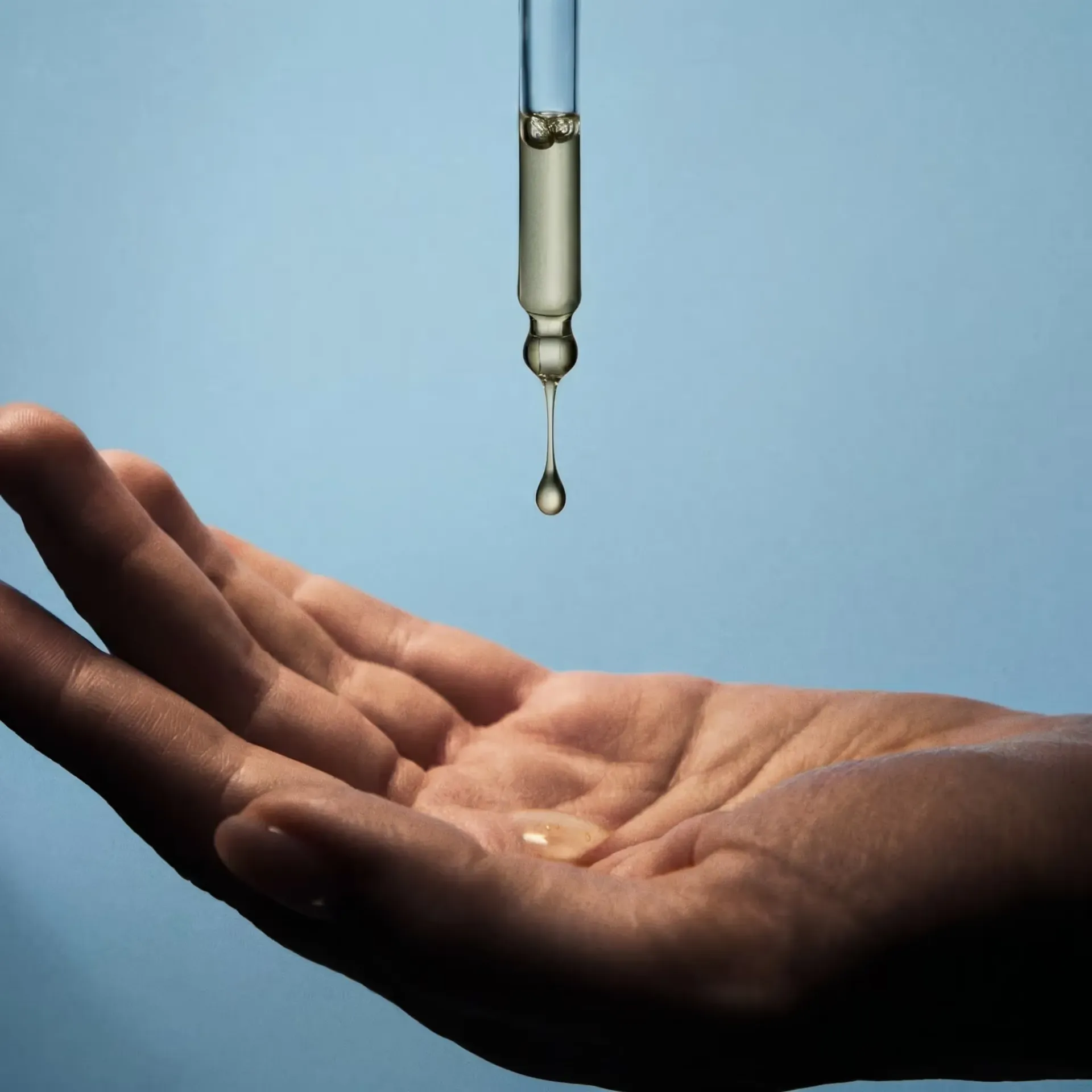 A glass dropper releases a golden oil drop onto an open palm against a blue background.