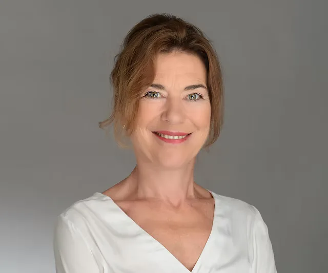 Professional portrait of a smiling woman with brown hair wearing a white blouse against a grey background.