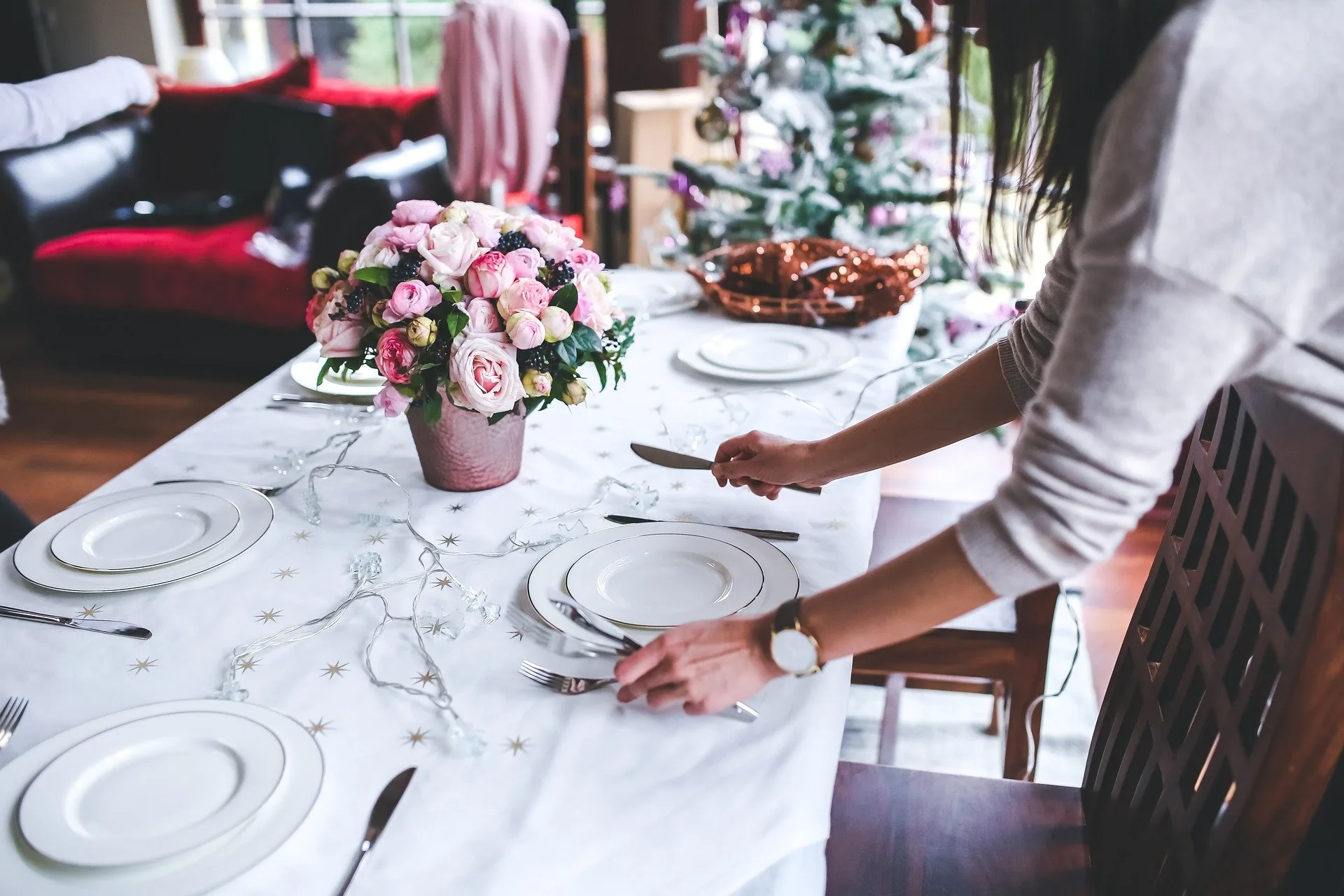 A woman sets an elegant Christmas dinner table with white plates, cutlery, and pink roses.