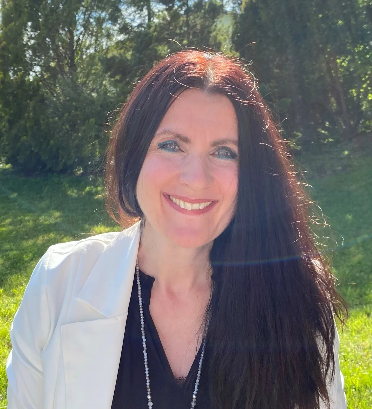 Smiling dark-haired woman in white blazer outdoors in sunlight, professional portrait of practice owner Anita Paul.
