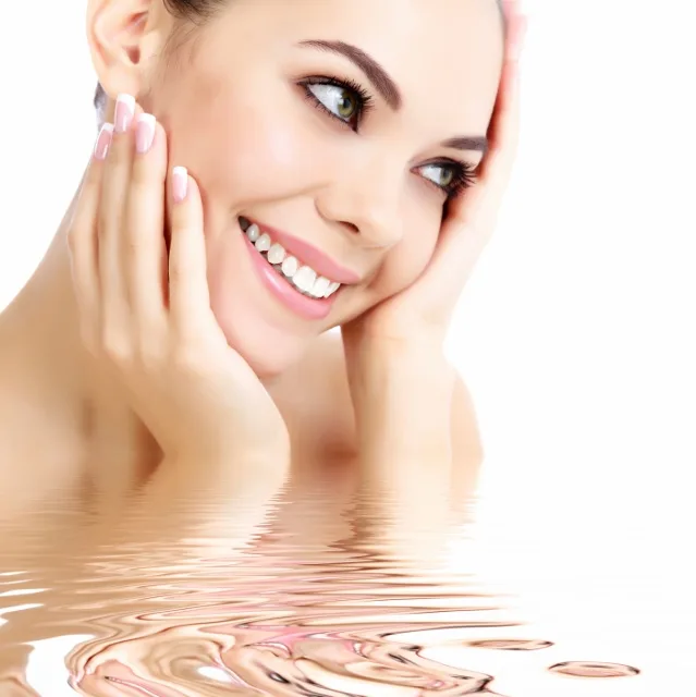 Smiling young woman with glowing skin resting her face gently in her hands near water.