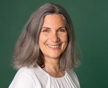 A smiling middle-aged woman with grey hair against a green background, conveying warmth and approachability.