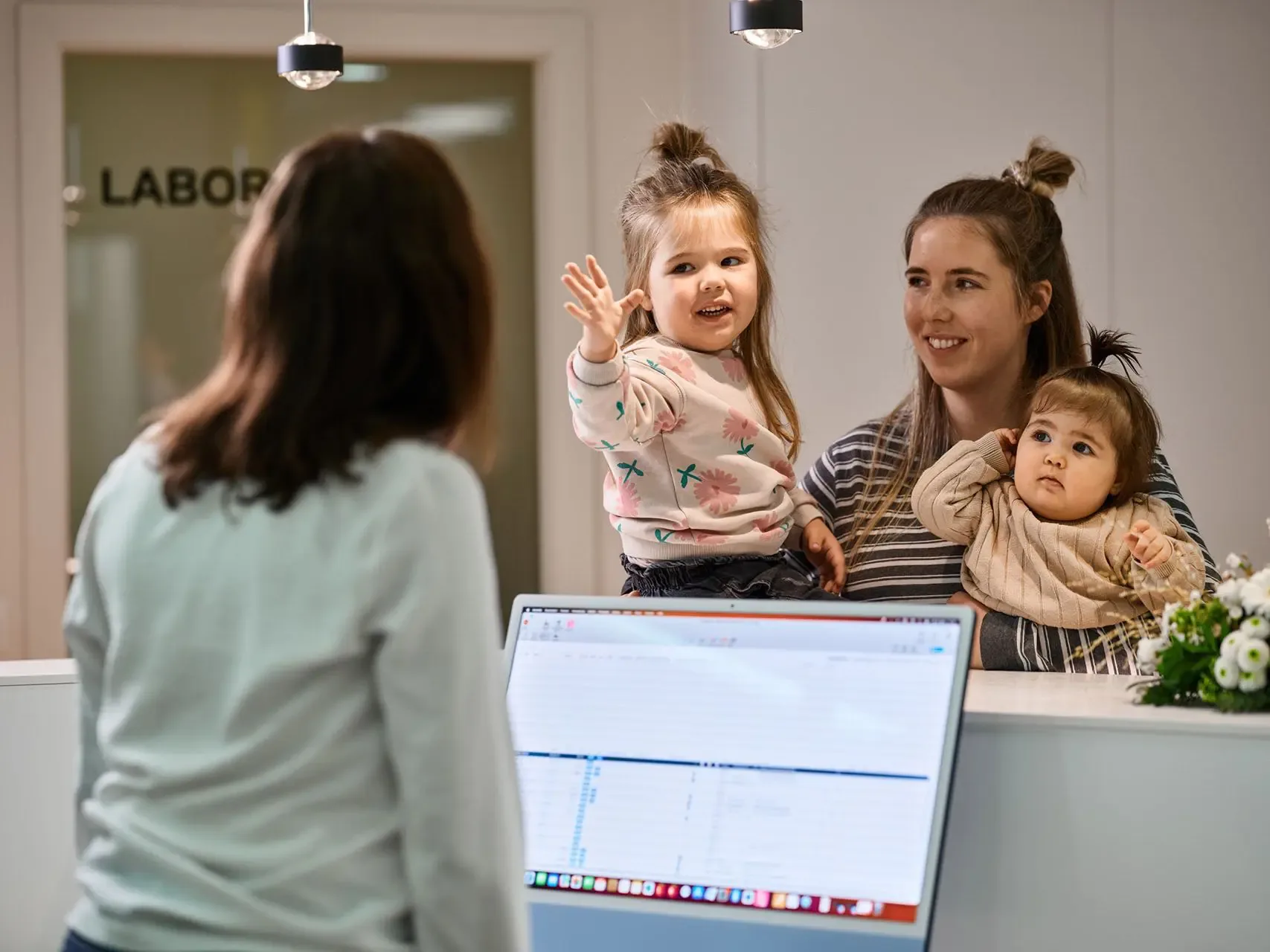 A young mother with two small children at the reception desk, illustrating the practice's welcoming, family-friendly atmosphere for all generations.