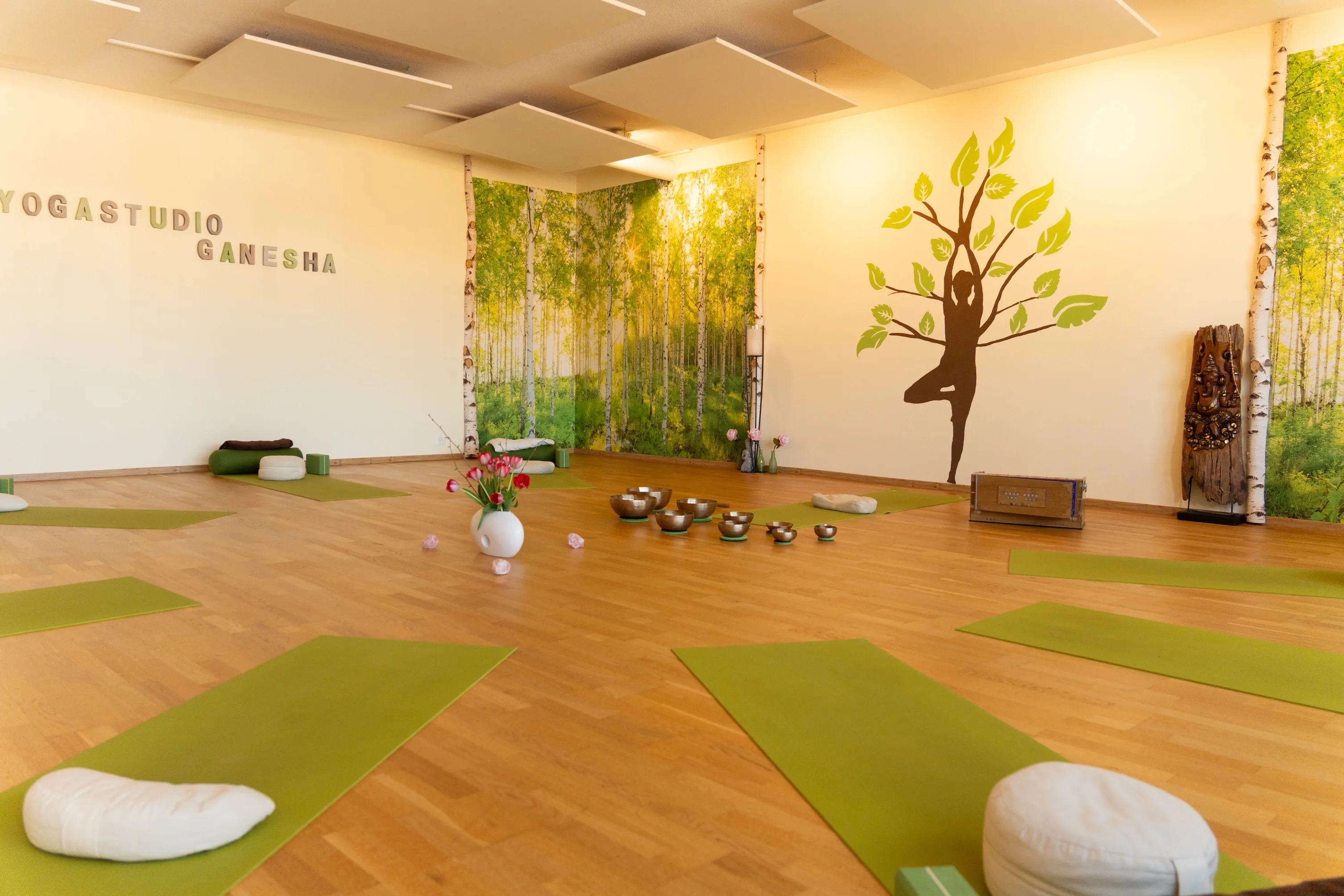 Spacious Yogastudio Ganesha hall with green mats, singing bowls, and a birch forest wallpaper.
