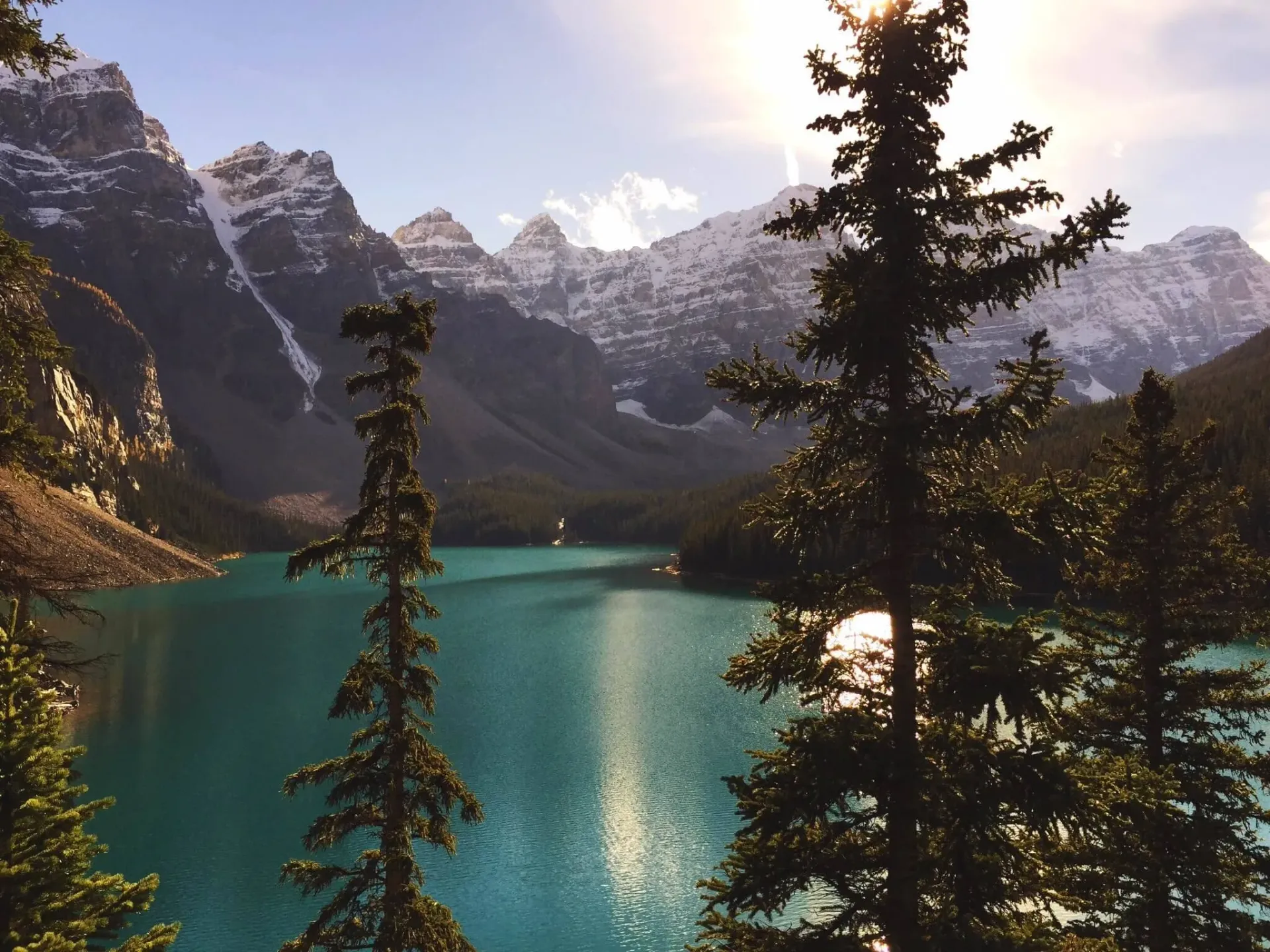 Turquoise glacial lake surrounded by snow-capped mountains and pine trees at golden hour.
