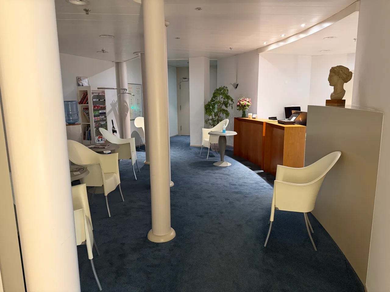 Spacious waiting area with cream chairs, blue carpet, reception desk, and classical bust sculpture.