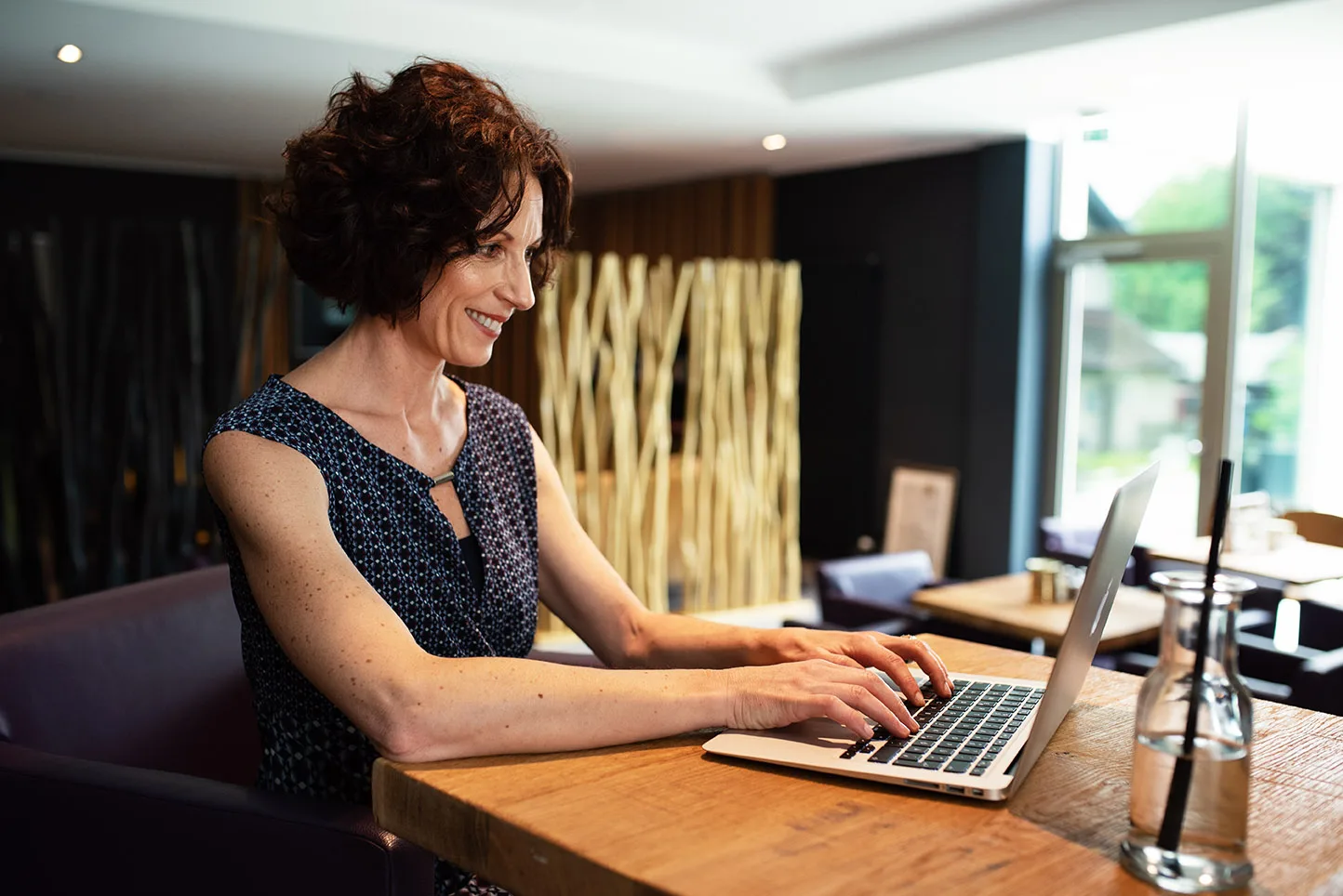 Claudine Hofer sits confidently at a laptop in a warm café setting, embodying the approachable, modern professional behind Atemraum Aarau.
