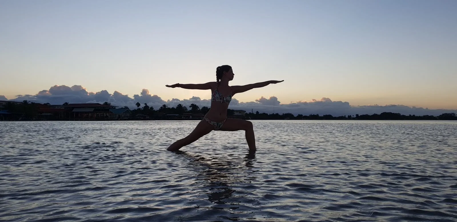 A woman holds warrior pose in shallow water at sunset with calm expansive horizon.