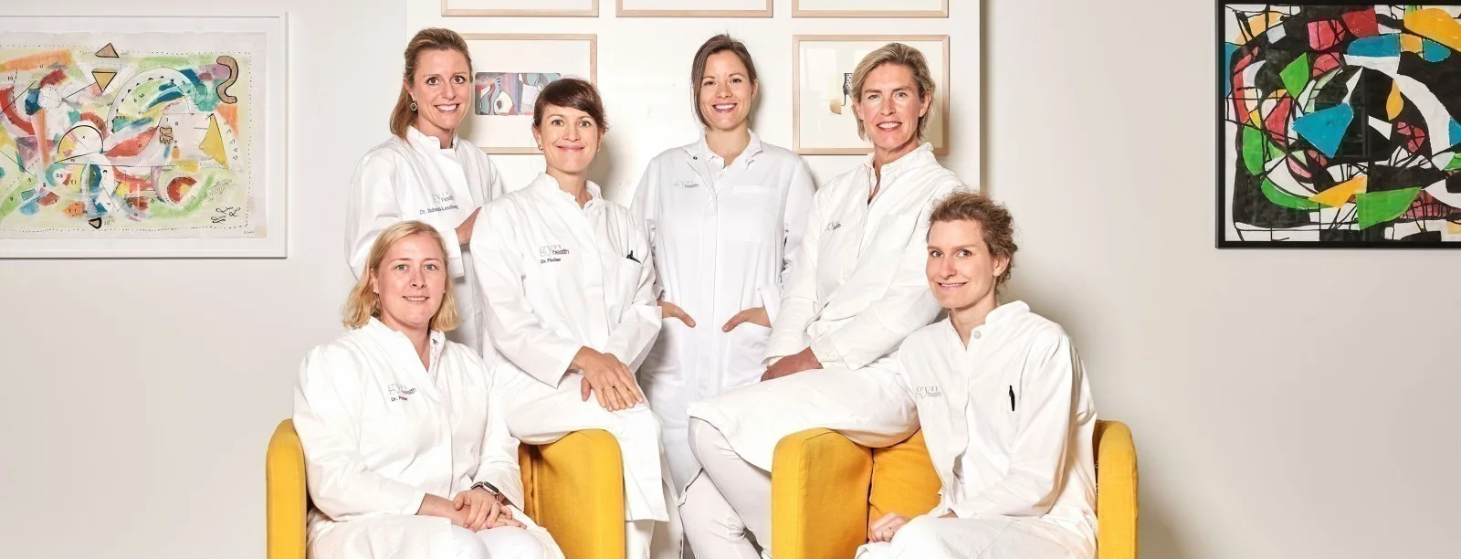 A large group of female doctors in white coats pose together on yellow chairs.