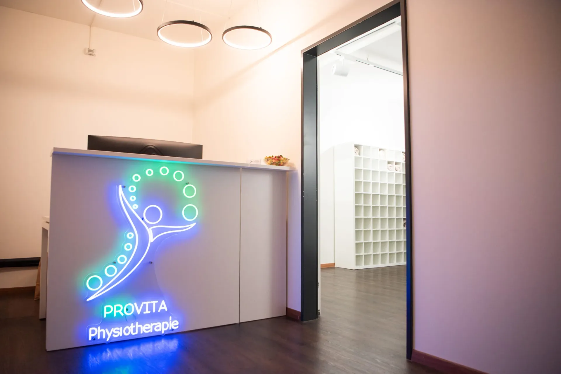 The glowing neon Provita Physiotherapie logo sign is displayed on the reception desk in the clinic entrance.