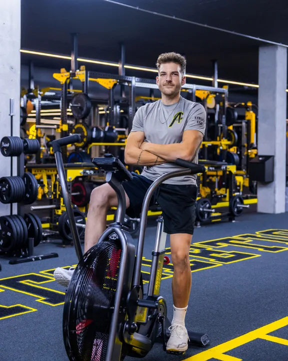 A confident trainer in Athletic Care branded shirt poses on an air bike, embodying the studio's ethos of purposeful, expert-guided training.