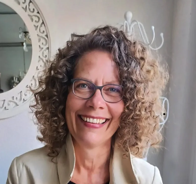 A smiling woman with curly hair and glasses poses warmly in a bright indoor setting.