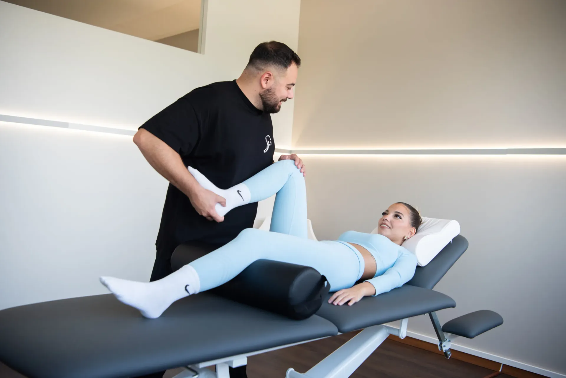 A physiotherapist in a black uniform smiles while performing a leg mobilization on a patient lying on a treatment table.