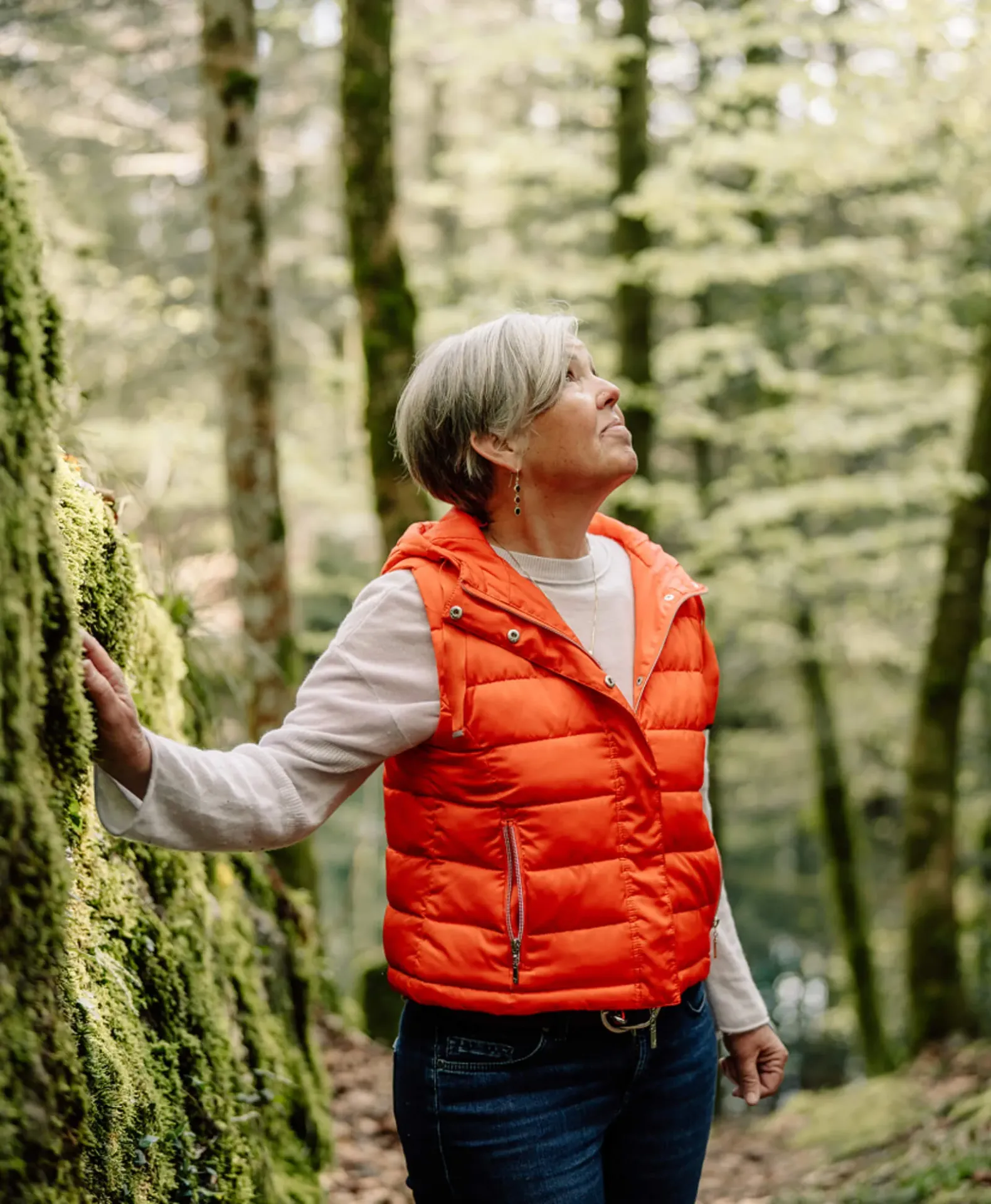 Woman in orange vest touches a moss-covered rock wall in a lush green forest.