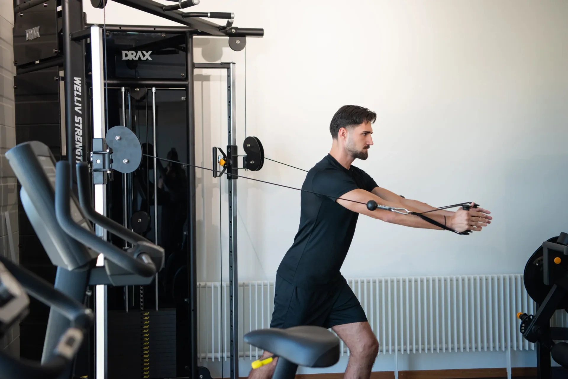 A male patient in black performs a cable pull exercise in a squat position on gym equipment.