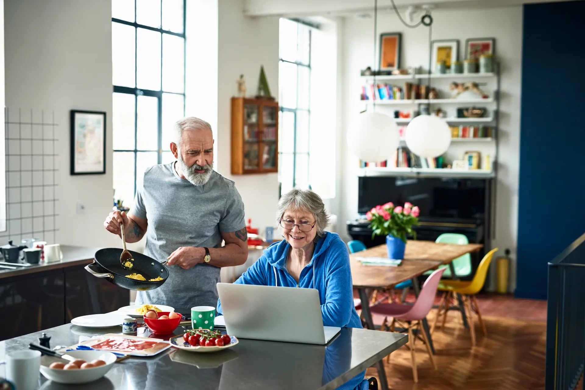 An older couple cooking and using a laptop together in a bright kitchen, illustrating how holistic nutrition guidance supports everyday life.