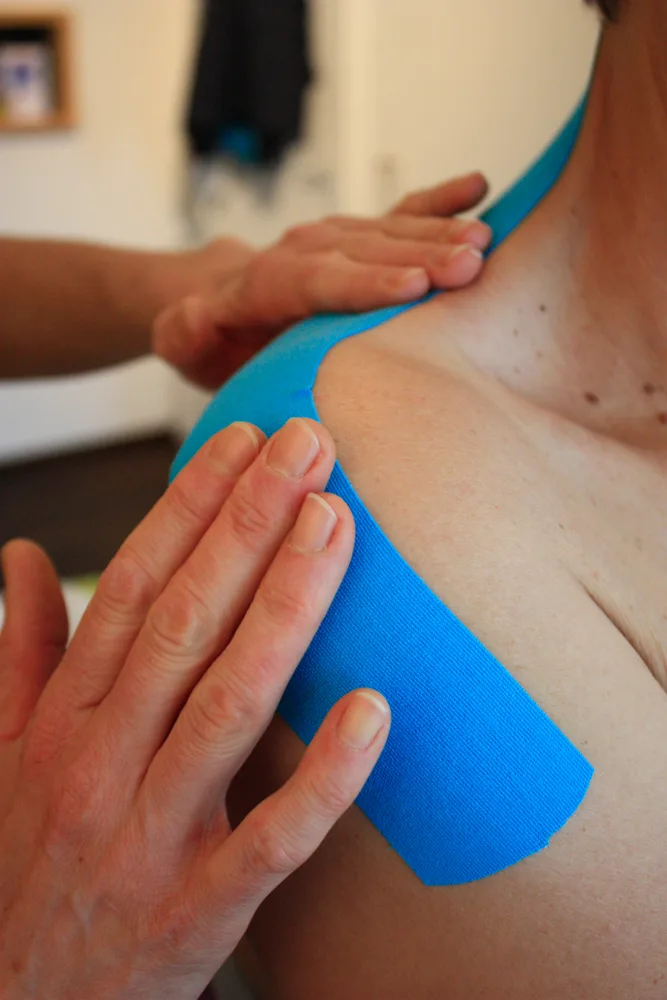 Therapist applies blue kinesiology tape to a client's shoulder area.