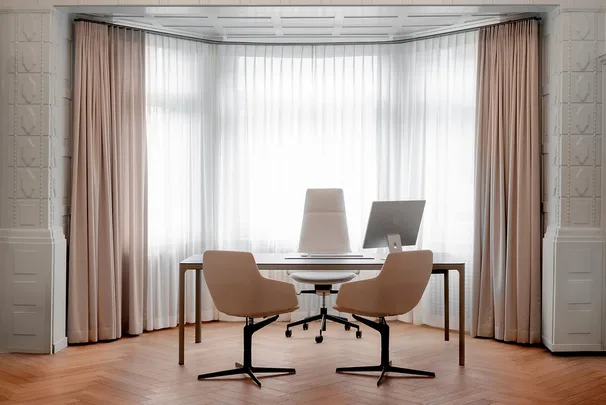 Consultation office with bay window, blush curtains, and two visitor chairs at desk.