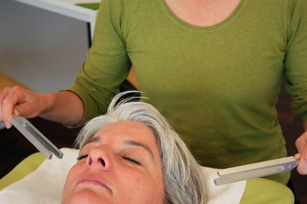 A therapist holds tuning forks beside a relaxed client lying on a treatment table.