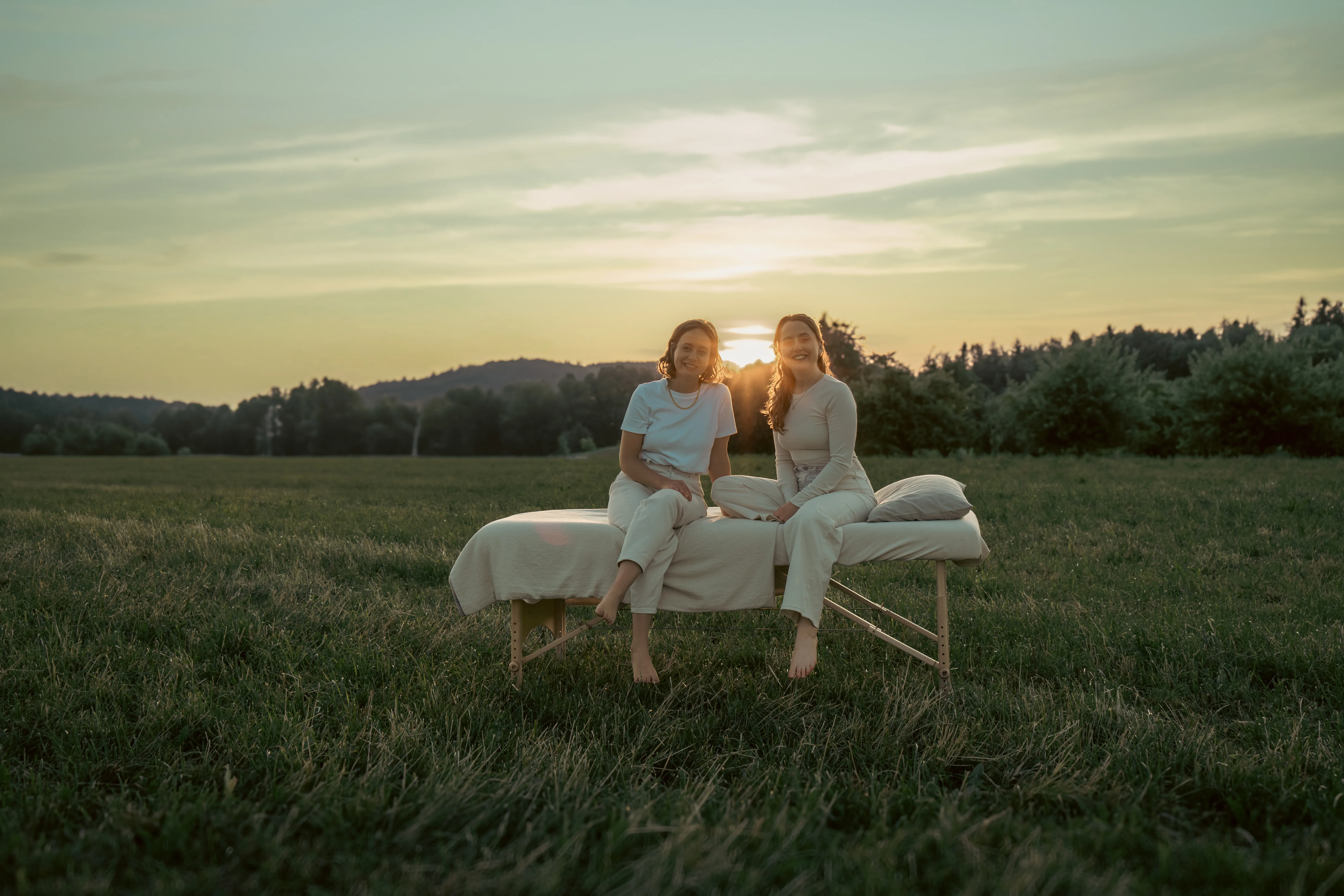 Two women sit together on an outdoor massage table in a sunlit green meadow at golden hour.