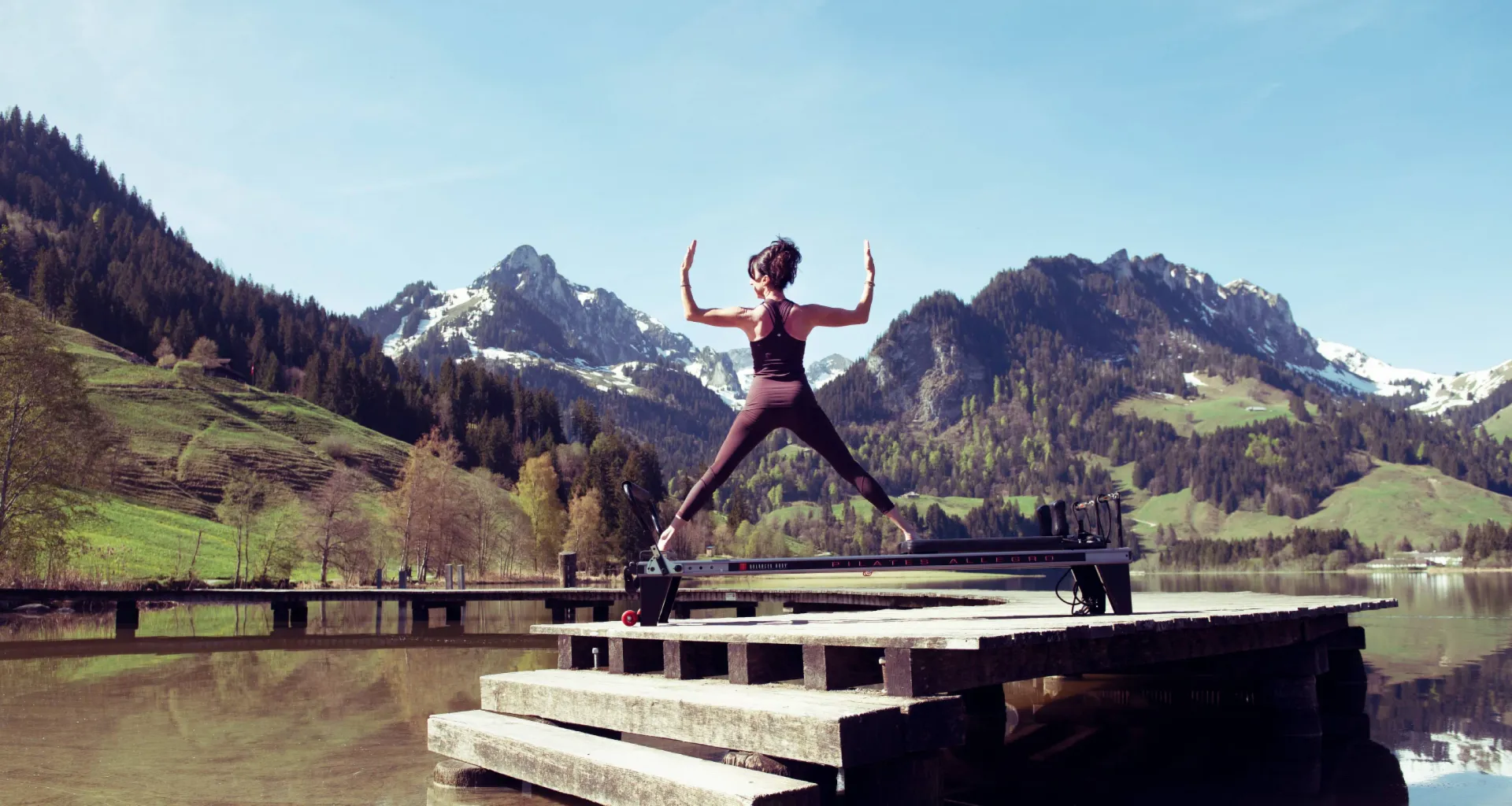 A woman stands in a wide-legged stance on a lakeside dock with snow-capped Alps behind her.