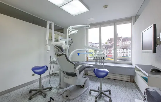 A clean, modern dental treatment room with a professional chair, blue stools, and city view.