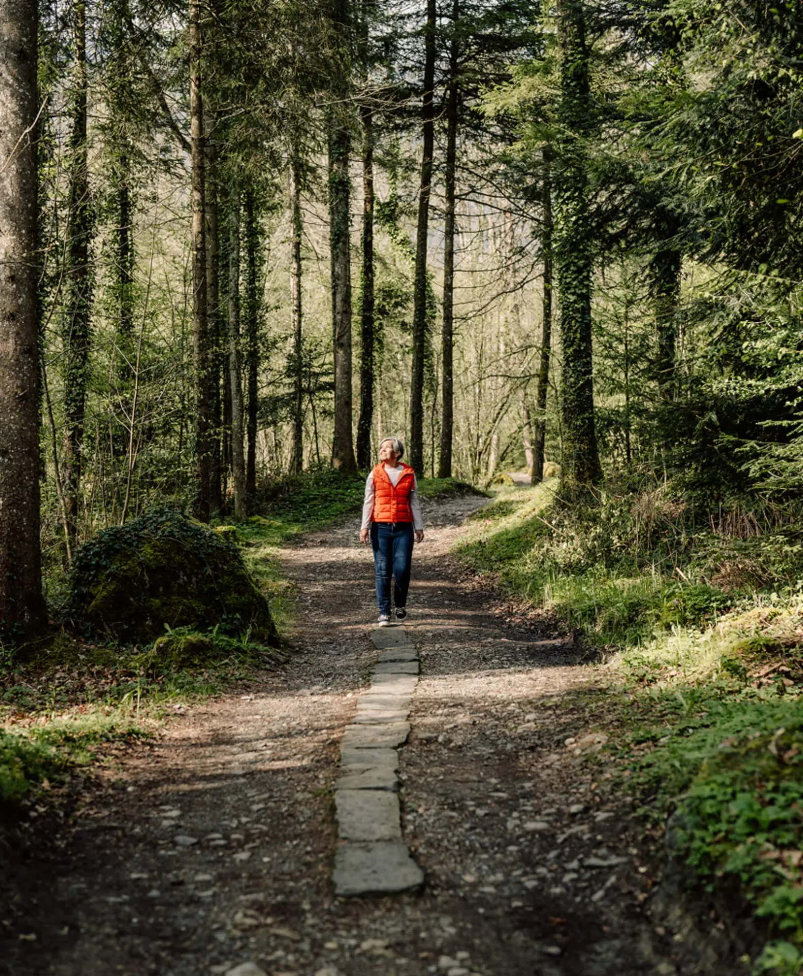 Woman in orange vest walks mindfully along a sunlit forest path.