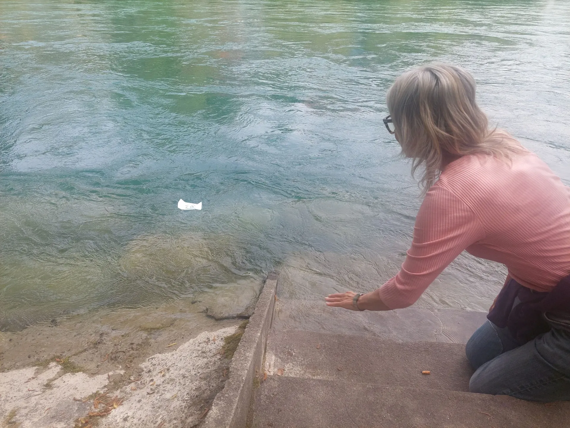 A woman watches a small paper boat float away on the turquoise river, a symbolic release.