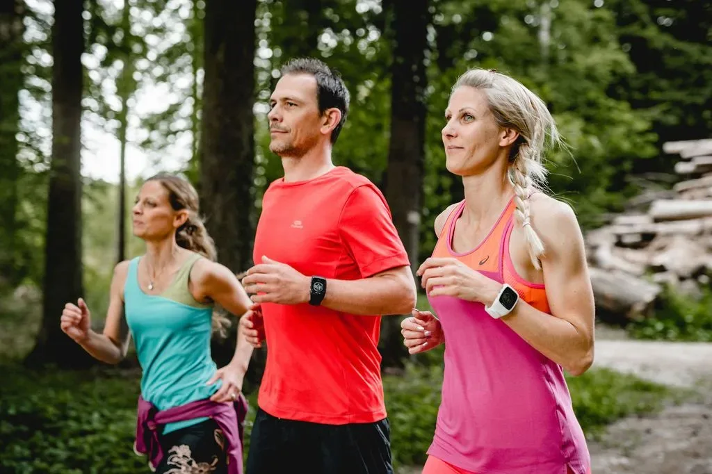 Three adults in colorful sportswear jogging together through a sunlit forest path.