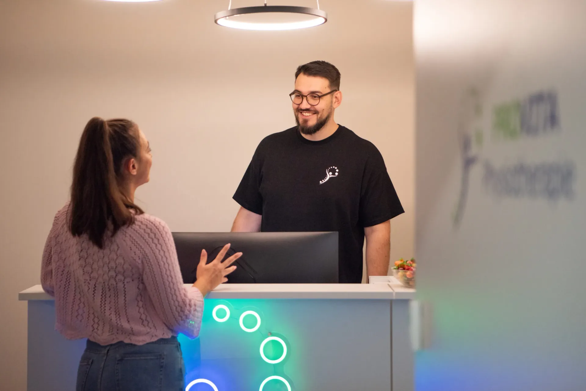 A smiling receptionist in a black shirt greets a female visitor at the illuminated Provita reception desk.