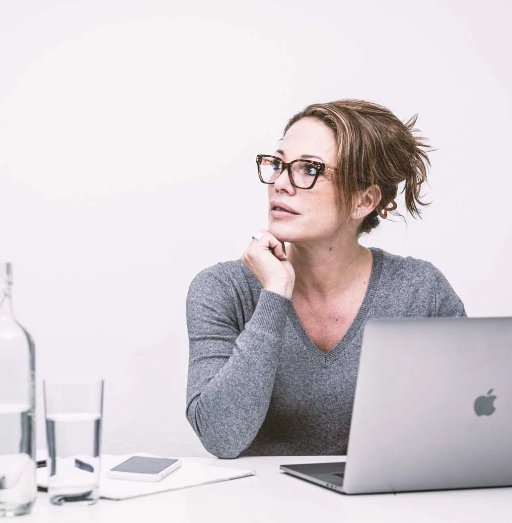 A thoughtful woman in glasses works at a laptop in a clean, professional setting.