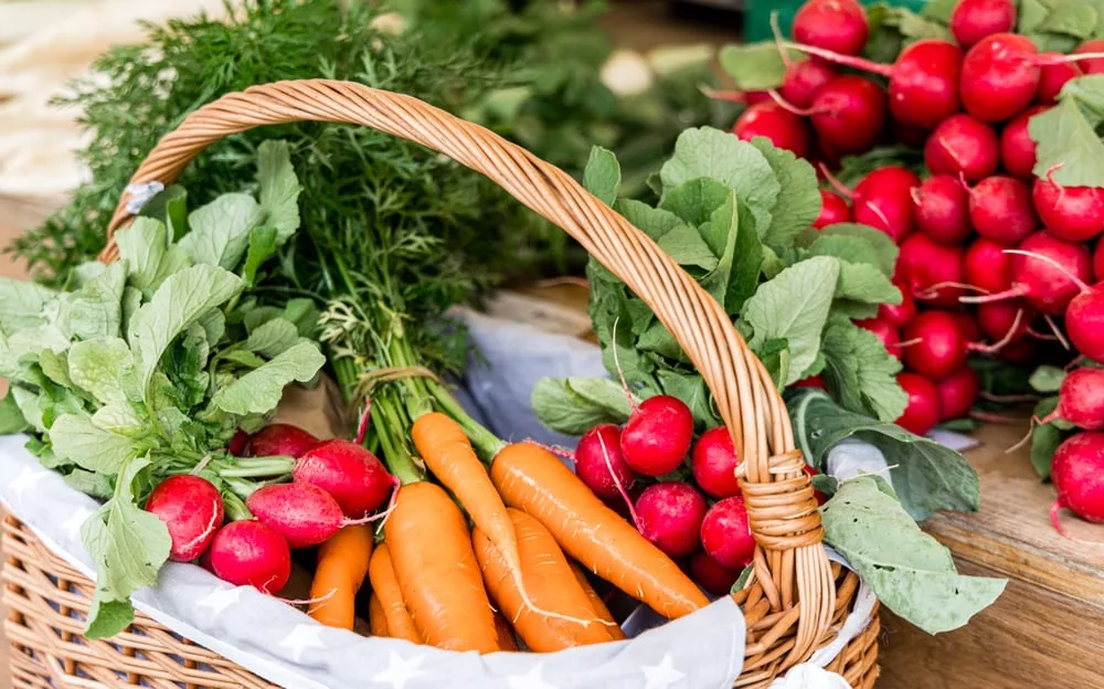 A wicker basket filled with fresh carrots and radishes, alongside a bunch of radishes on a wooden surface.