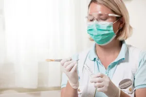 A female dental professional in scrubs and mask carefully inspects a dental instrument.