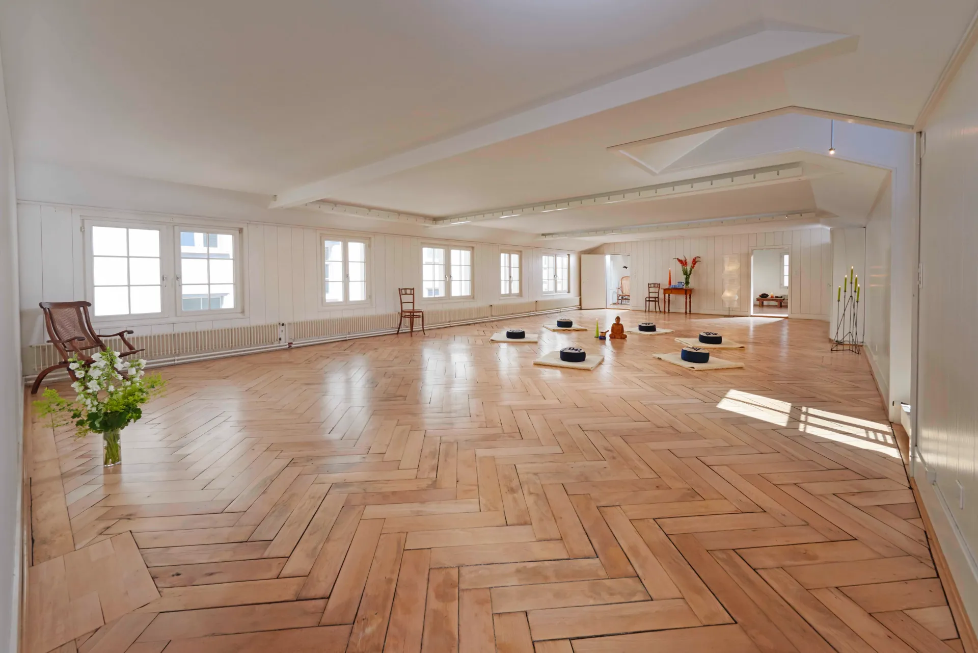 Spacious white studio viewed from the side, showing cushions, a Buddha figurine, and tall windows.