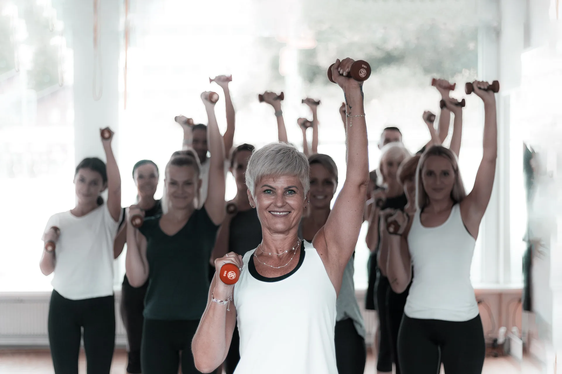 A silver-haired instructor leads a joyful group dumbbell class, perfectly capturing FIT-N-FUN's community spirit and personal training philosophy.