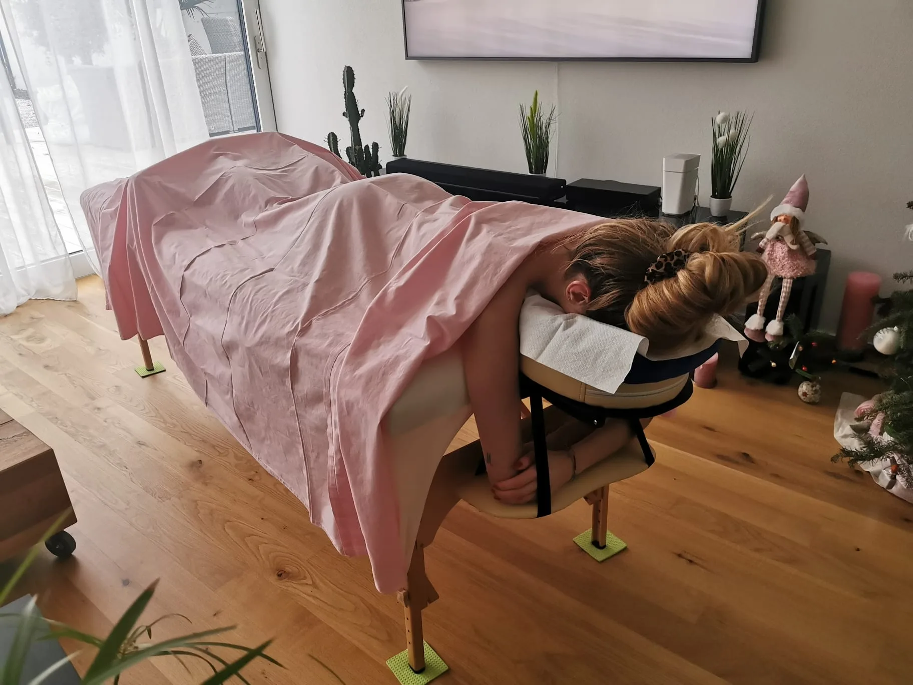 A client rests face-down on a massage chair draped in pink fabric in a bright home setting.