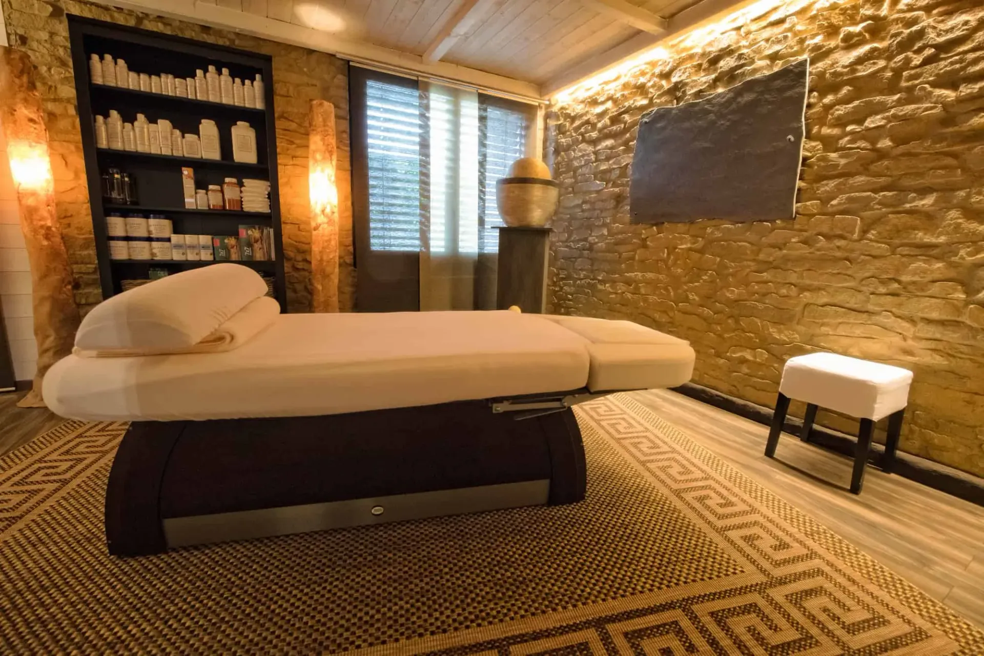 Smiling therapist performs a facial treatment with golden tools on a client in a rustic stone-walled room.